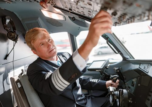 Male airline captain holding control wheel and turning switch on overhead panel while sitting in airplane cockpit
